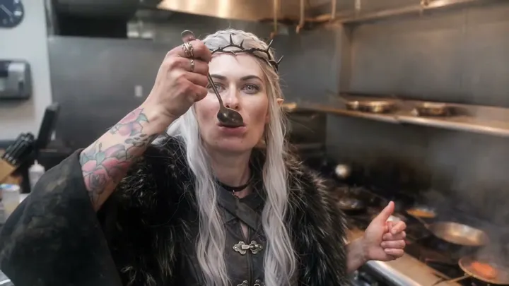 An elf queen in a fur mantle steps into the kitchen and tastes the sauce in the chef’s place.