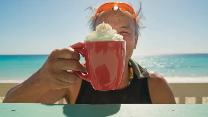A seaside sip keeps its motion and outfit while a new laughing face takes over.